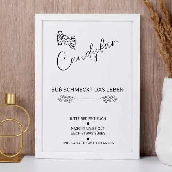 Ein gerahmtes Schild mit der Aufschrift Candybar in deutscher Sprache, verziert mit einer kleinen Süßigkeitengrafik, steht auf einer hölzernen Fläche neben einem goldenen Kerzenhalter und getrockneten Gräsern. Das Schild lädt die Gäste ein, Süßigkeiten zu genießen.