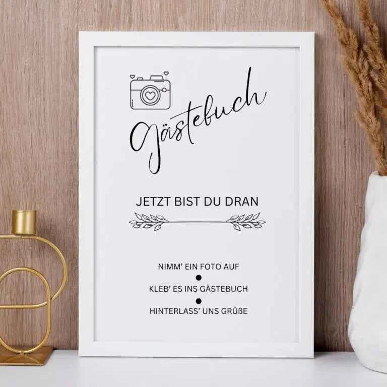 Gerahmtes deutsches Schild auf einer hölzernen Oberfläche mit dekorativen Pflanzen und einem Kerzenhalter in der Nähe. Das Schild fordert die Gäste auf, ein Foto zu machen, es in das Gästebuch zu kleben und einen Gruß zu hinterlassen.