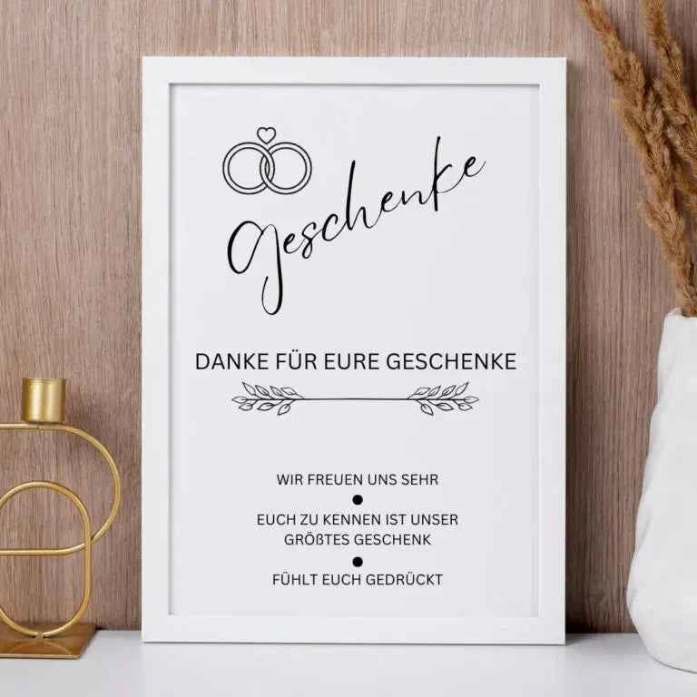 Ein gerahmtes Schild mit Eheringen und dem Wort "Geschenke". Darunter steht ein deutscher Text, der den Gästen für ihre Geschenke dankt und zum Ausdruck bringt, dass es das größte Geschenk ist, sie zu kennen. Der Rahmen steht auf einem Schrank neben einem Kerzenhalter aus Messing und getrocknetem Gras.