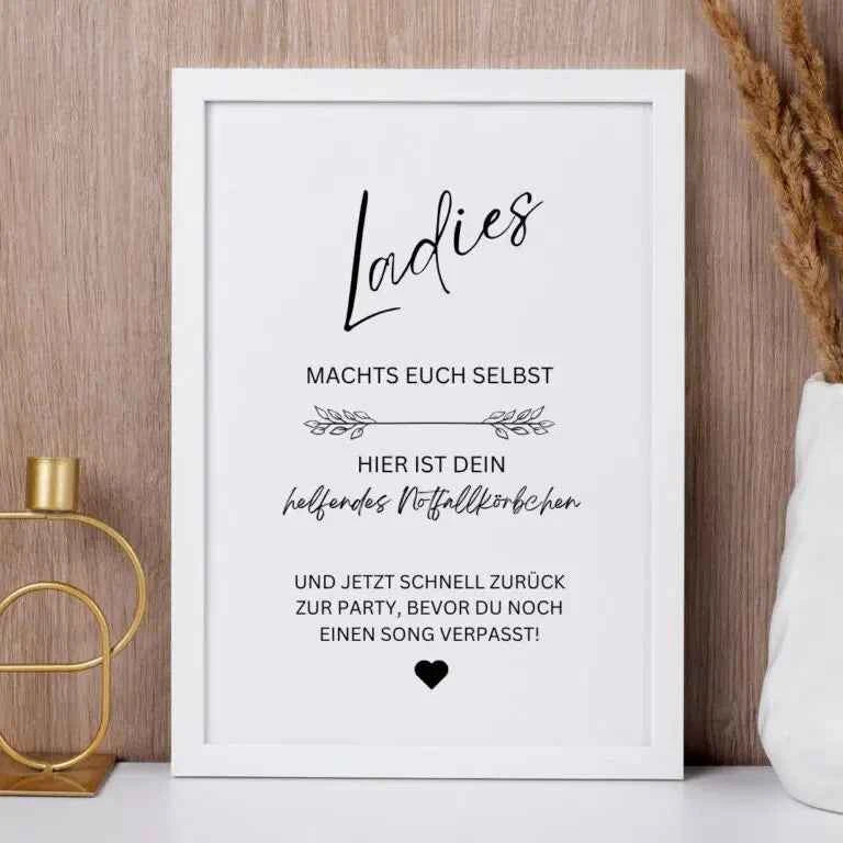 Ein gerahmtes deutsches Schild auf einem weißen Regal lautet: Meine Damen, macht euch selbst. Hier ist dein helfendes Notfallkörbchen. Und jetzt schnell zurück zur Party, bevor du noch einen Song verpasst!.