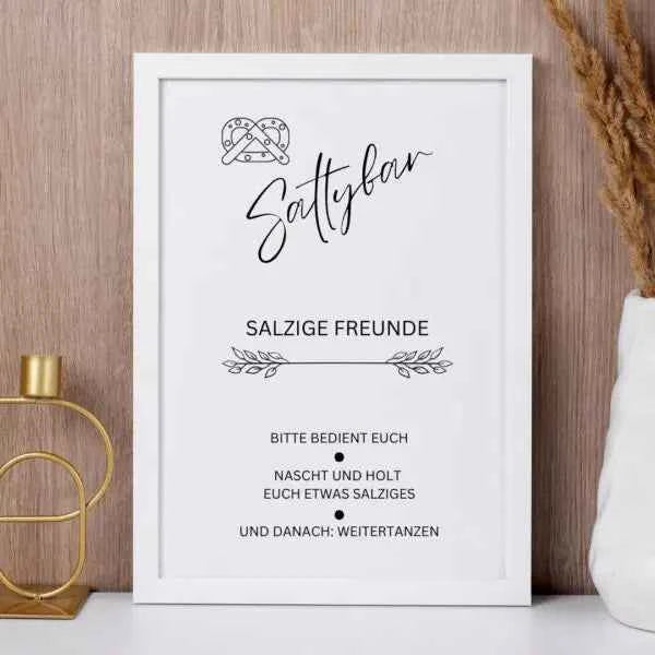 Ein gerahmtes Schild auf einem Regal mit der Aufschrift Saltybar, Salzige Freunde. Bitte bedient euch, nascht und holt euch etwas Salziges und danach: Weitertanzen mit einer Brezelillustration über dem Text. Getrocknete Pflanzen und eine Kerze stehen daneben.