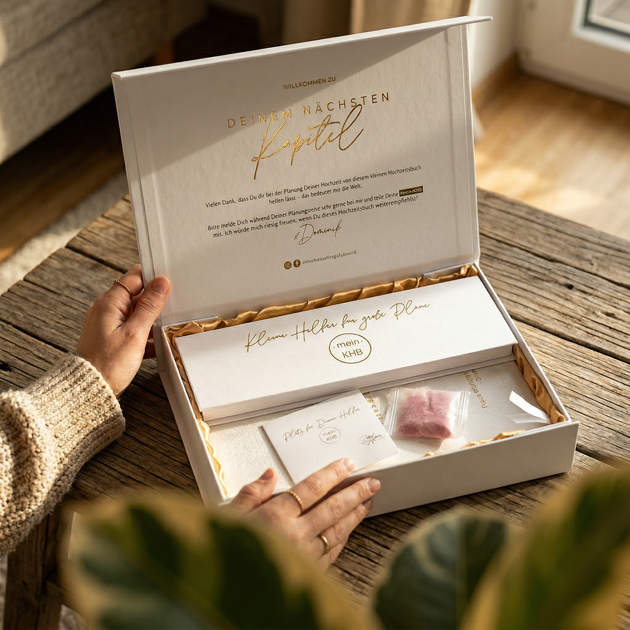 Eine Person öffnet eine weiße Geschenkschachtel mit einem Umschlag, einer Karte und einem kleinen Tütchen. Auf dem Deckel der Schachtel stehen ein goldener deutscher Text und ein Willkommensgruß. Die Szene ist auf einem Holztisch in der Nähe eines Fensters mit Pflanzen dargestellt.