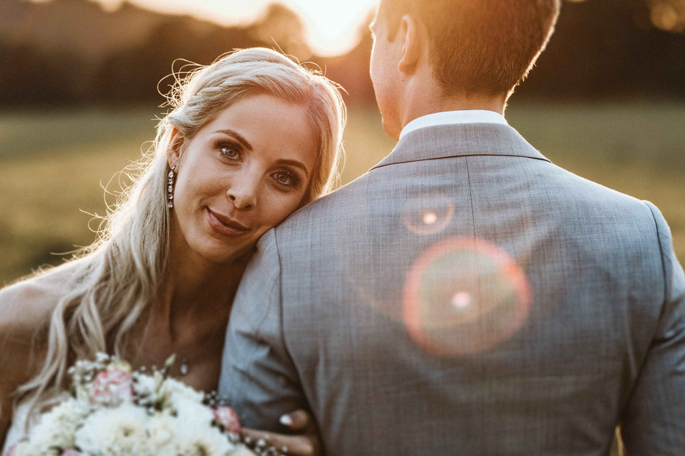 Eine blonde Frau im Hochzeitskleid lehnt ihren Kopf an die Schulter eines Mannes, der einen Blumenstrauß in der Hand hält, während das warme Sonnenlicht hinter ihnen in einer Freiluftumgebung aufblitzt.