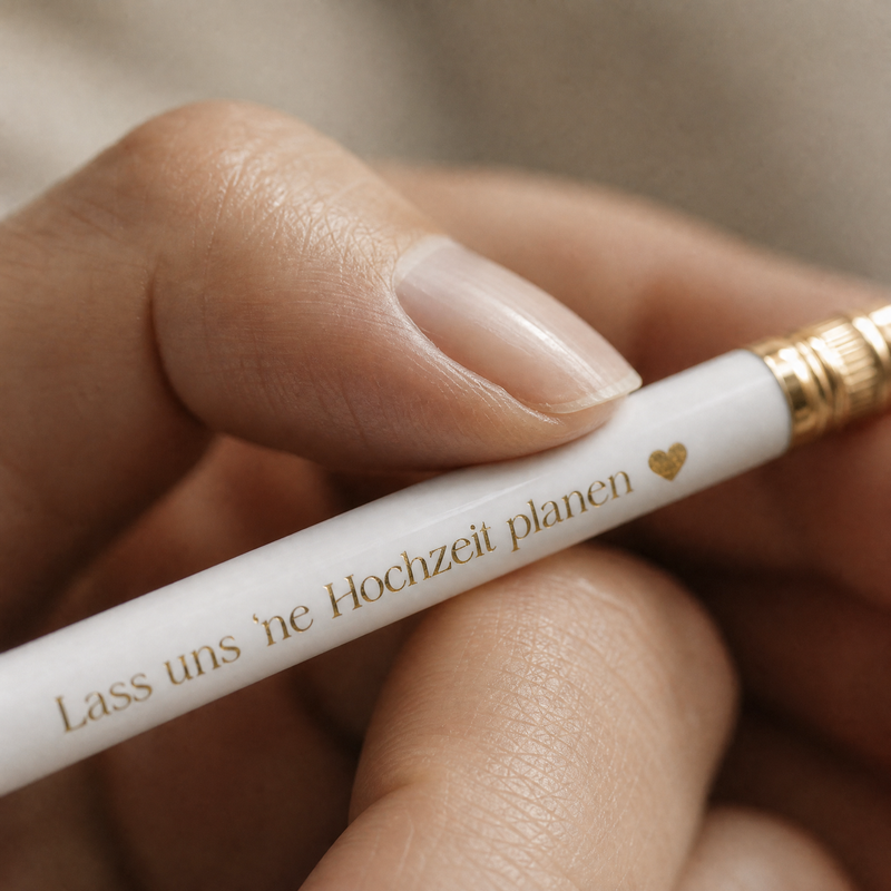 Eine Hand hält einen weißen Bleistift mit goldener Umrandung und deutschem Text, der lautet Lass uns eine Hochzeit planen, gefolgt von einem kleinen goldenen Herz.