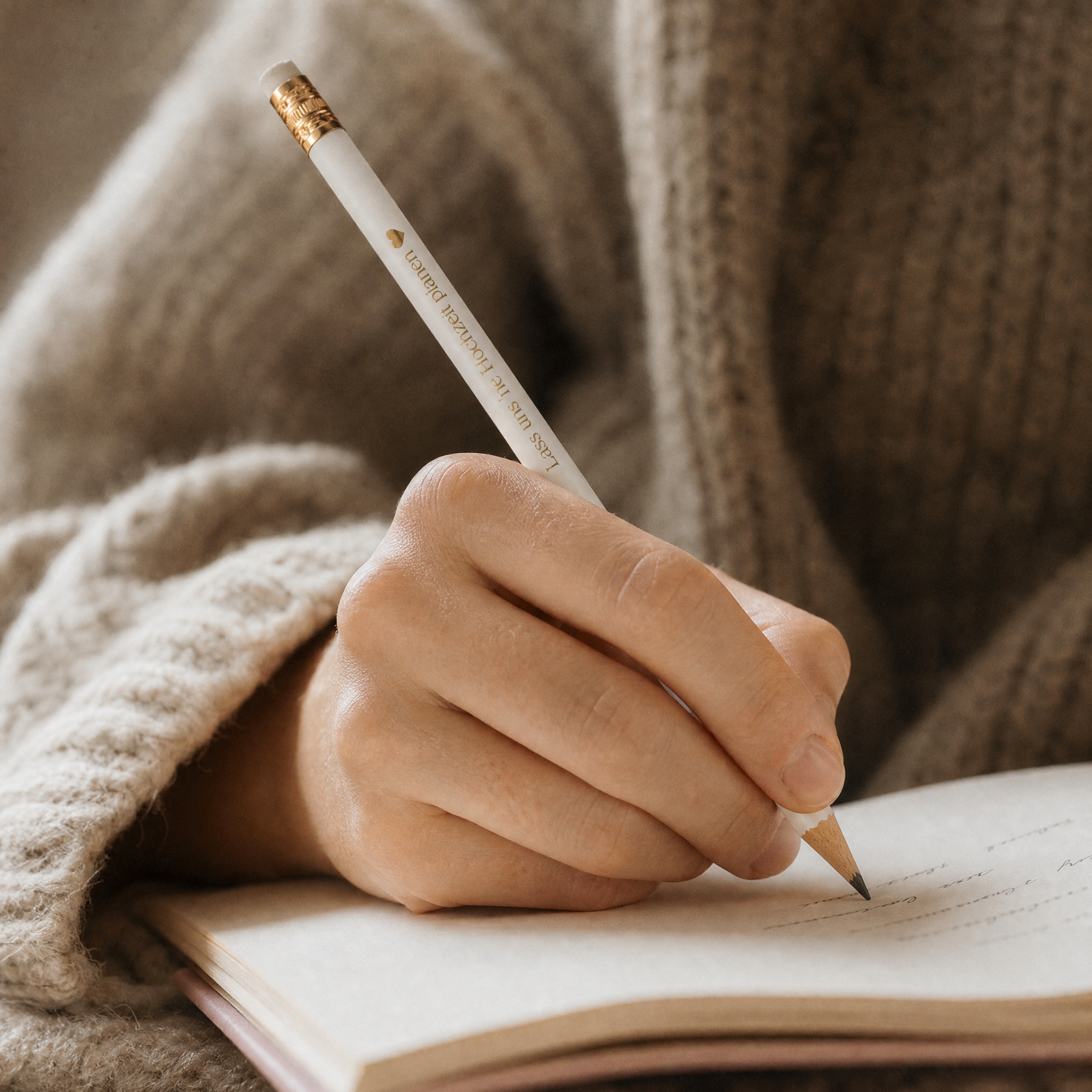 Eine Person, die einen gemütlichen, beigen Pullover trägt, schreibt mit einem weißen Bleistift in ein Notizbuch. Das Bild konzentriert sich auf die Hand, die den Bleistift hält, und das geöffnete Notizbuch.