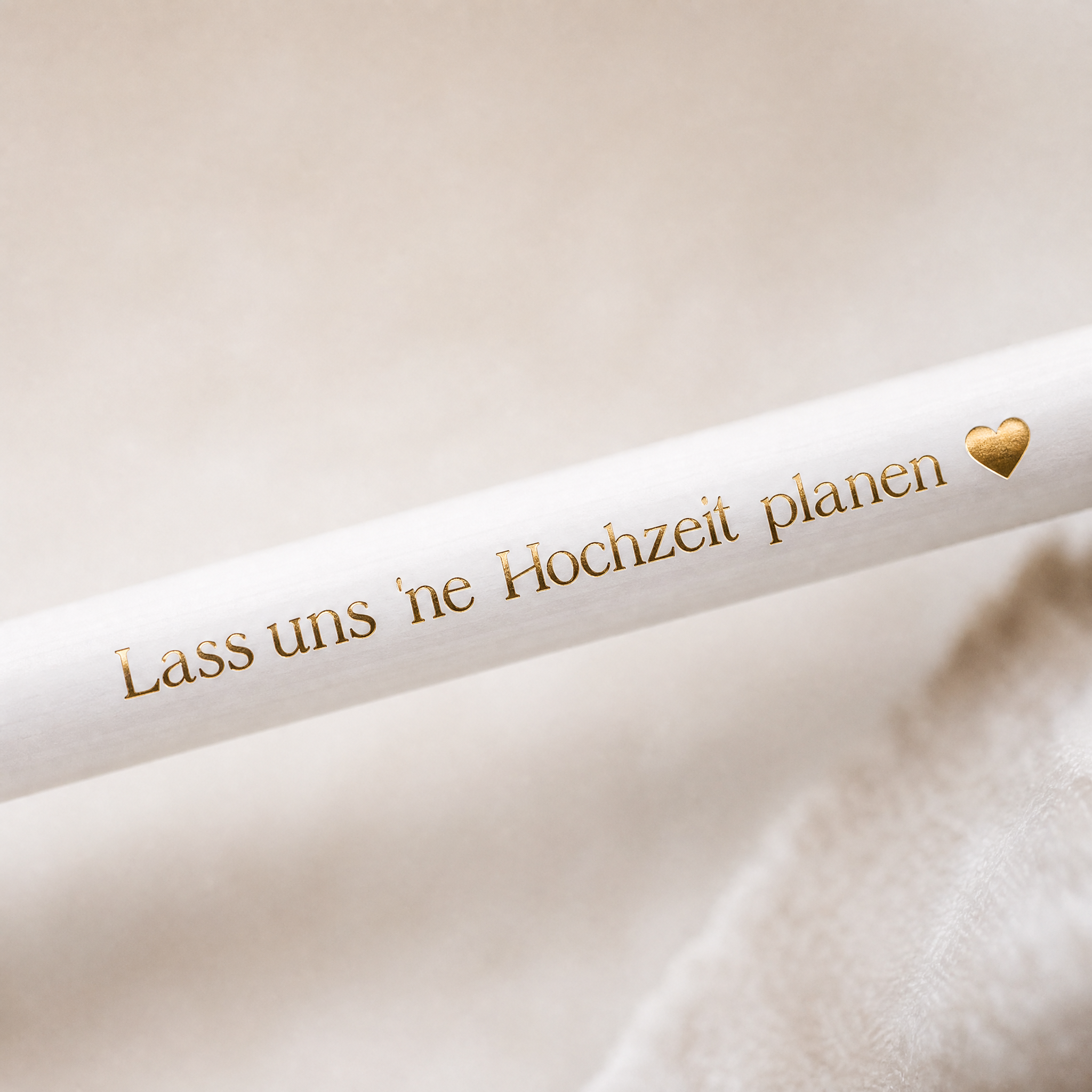 Ein weißer Stift mit goldener Schrift "Lass uns ne Hochzeit planen" und einem goldenen Herzsymbol. Der Hintergrund ist weich und beige.
