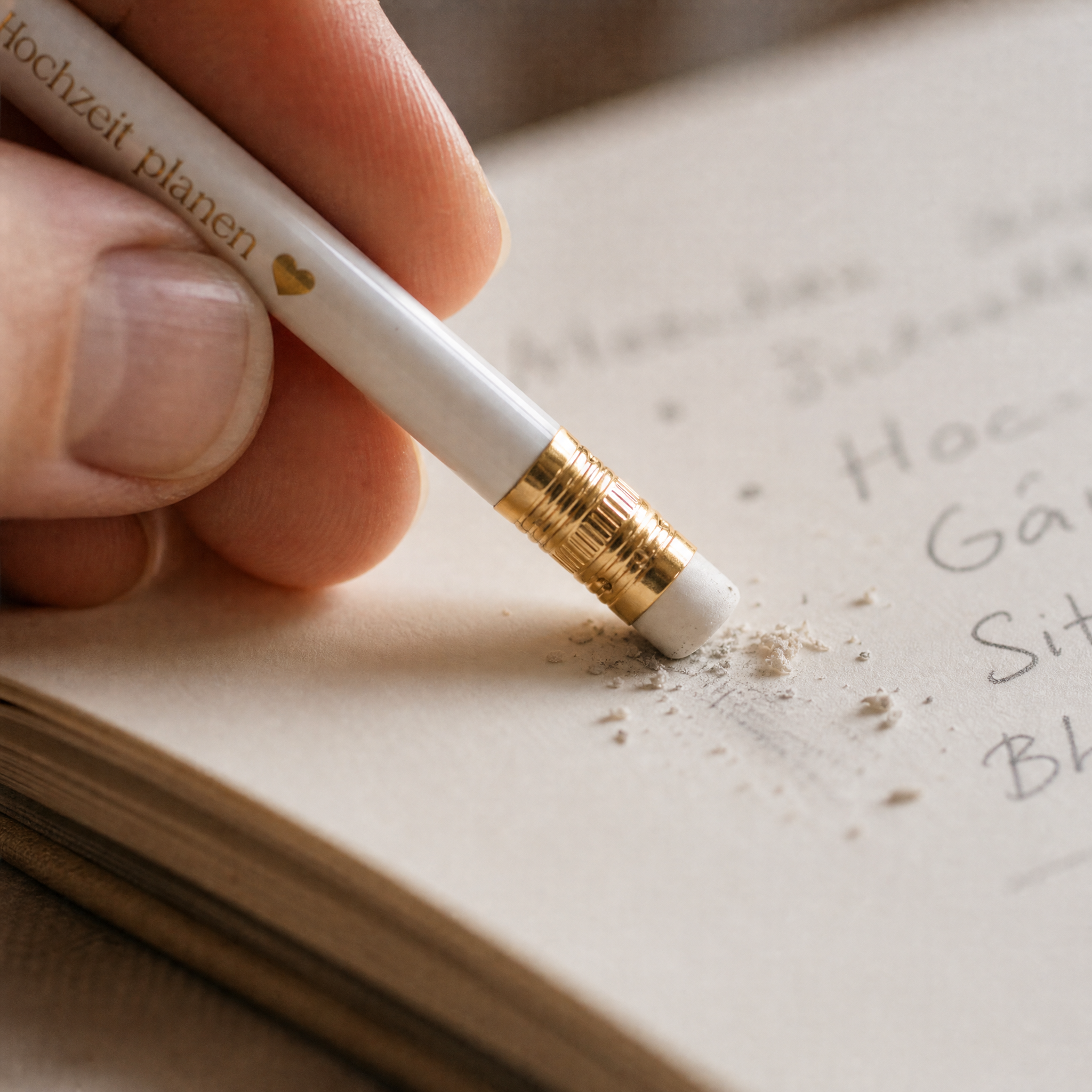 Nahaufnahme einer Hand, die mit einem weißen Bleistift mit goldenem Radiergummi auf Deutsch geschriebene Wörter aus einem Notizbuch löscht, wobei Radiergummisplitter auf dem Papier sichtbar sind.