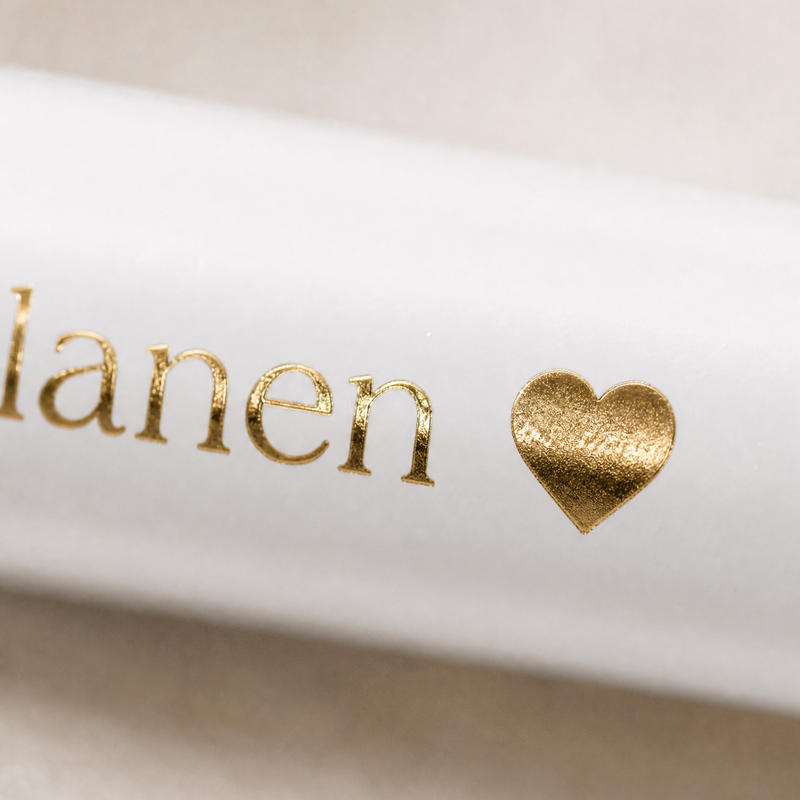 Eine Nahaufnahme von weißem Papier mit dem teilweise aus Goldfolie bestehenden Text "lanen" und einem aus Goldfolie bestehenden Herzsymbol daneben.