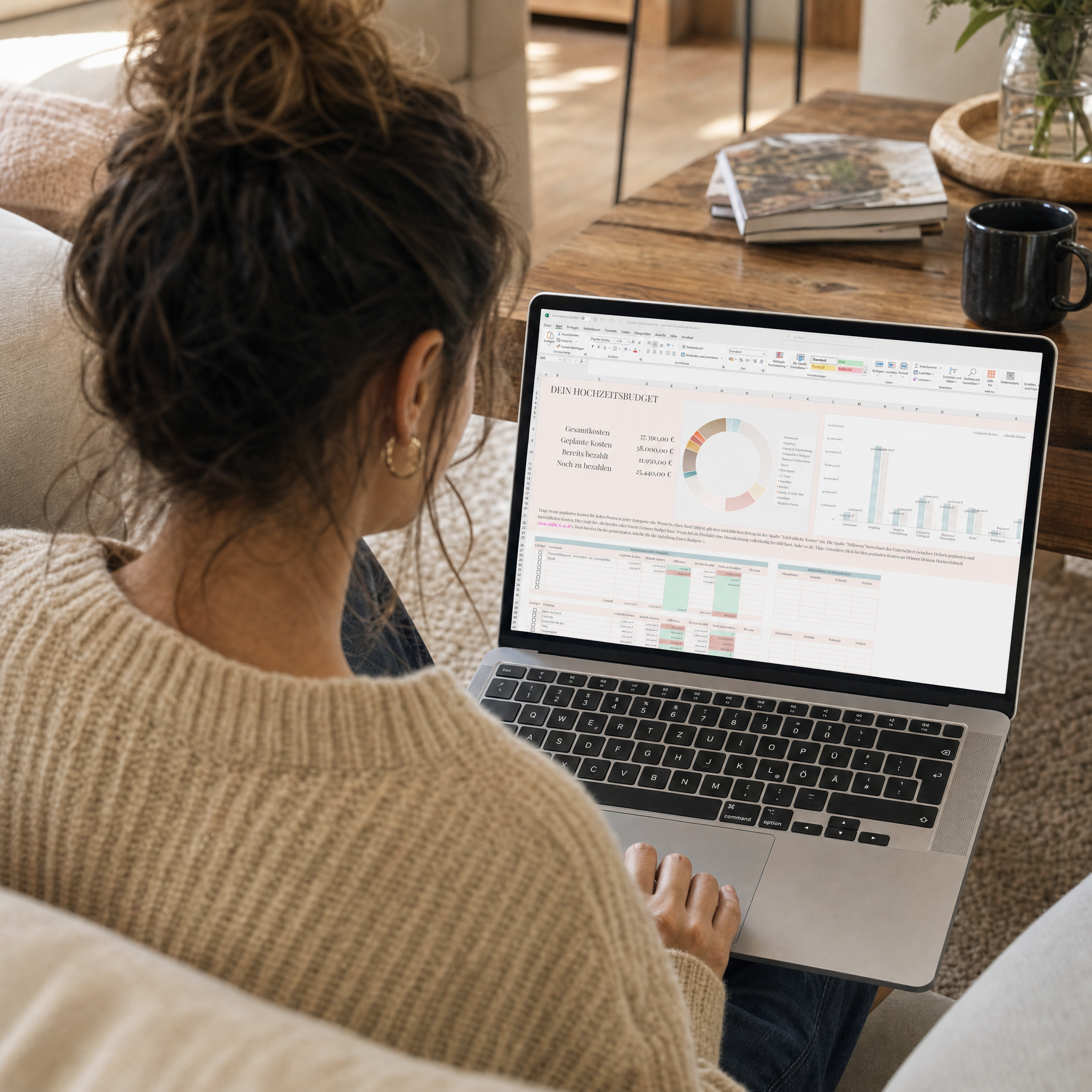Eine Frau mit lockigem Haar in einem Dutt sitzt auf einer Couch und arbeitet an einem Laptop, auf dem bunte Diagramme und Tabellen zu sehen sind. Eine Tasse und Bücher stehen auf dem Holztisch neben ihr in einem gemütlichen Wohnzimmer.