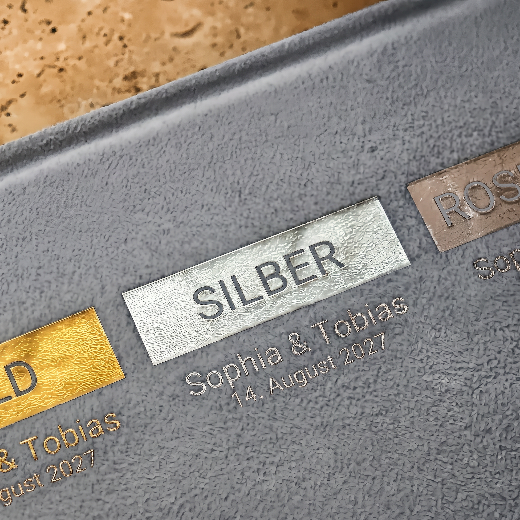 Auf einer strukturierten grauen Oberfläche befinden sich gravierte Metalltafeln mit den Bezeichnungen GOLD, SILBER und ROSE, unter denen Namen und das Datum 14. August 2027 sind unter jeder Tafel eingraviert.