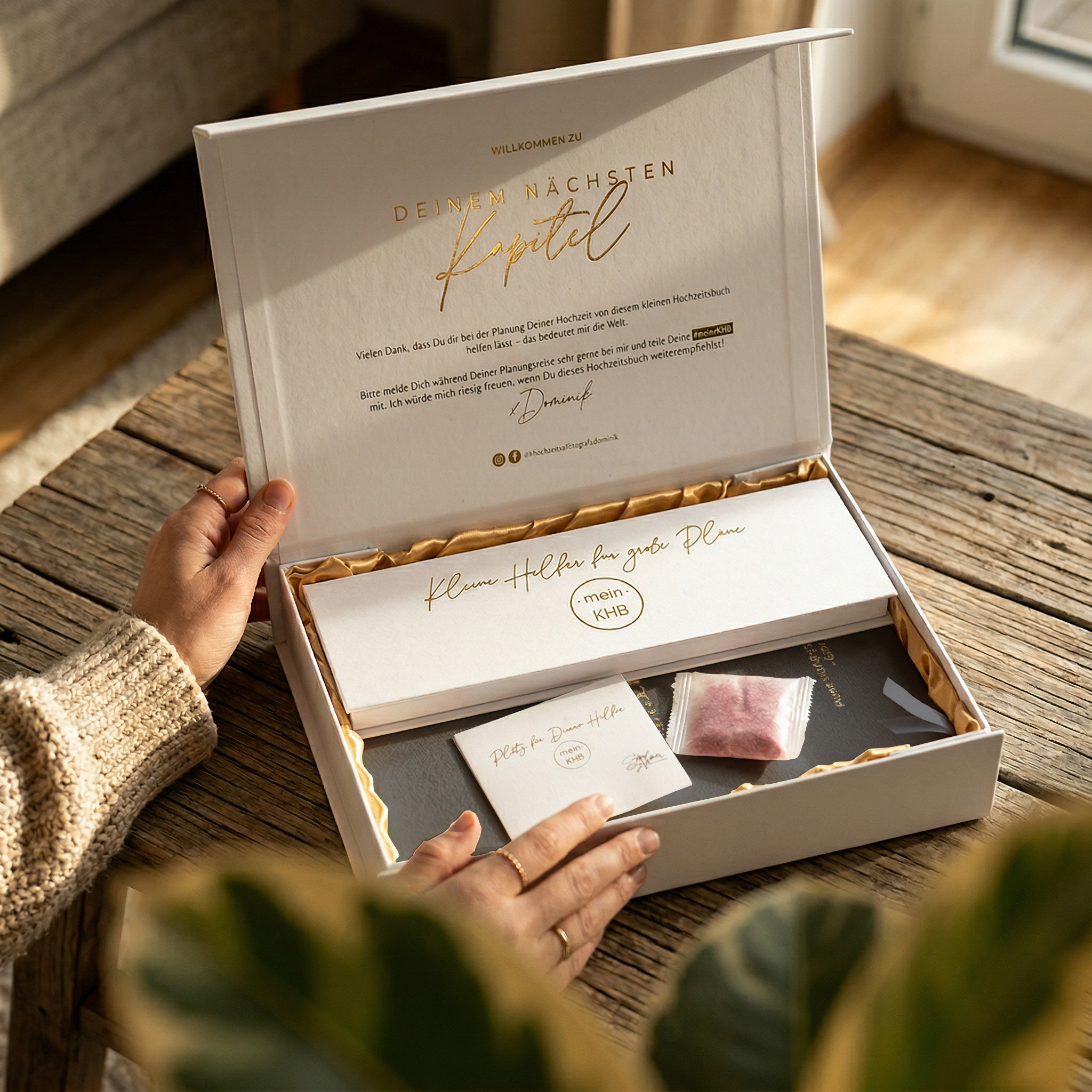 Eine Person öffnet eine weiße Geschenkschachtel auf einem Tisch. Die Schachtel enthält eine elegante Karte, einen kleineren Umschlag, ein Tütchen mit rosa Puder und Papierkonfetti. Der Deckel der Schachtel ist mit goldener und schwarzer deutscher Schrift und einer handschriftlichen Unterschrift versehen.