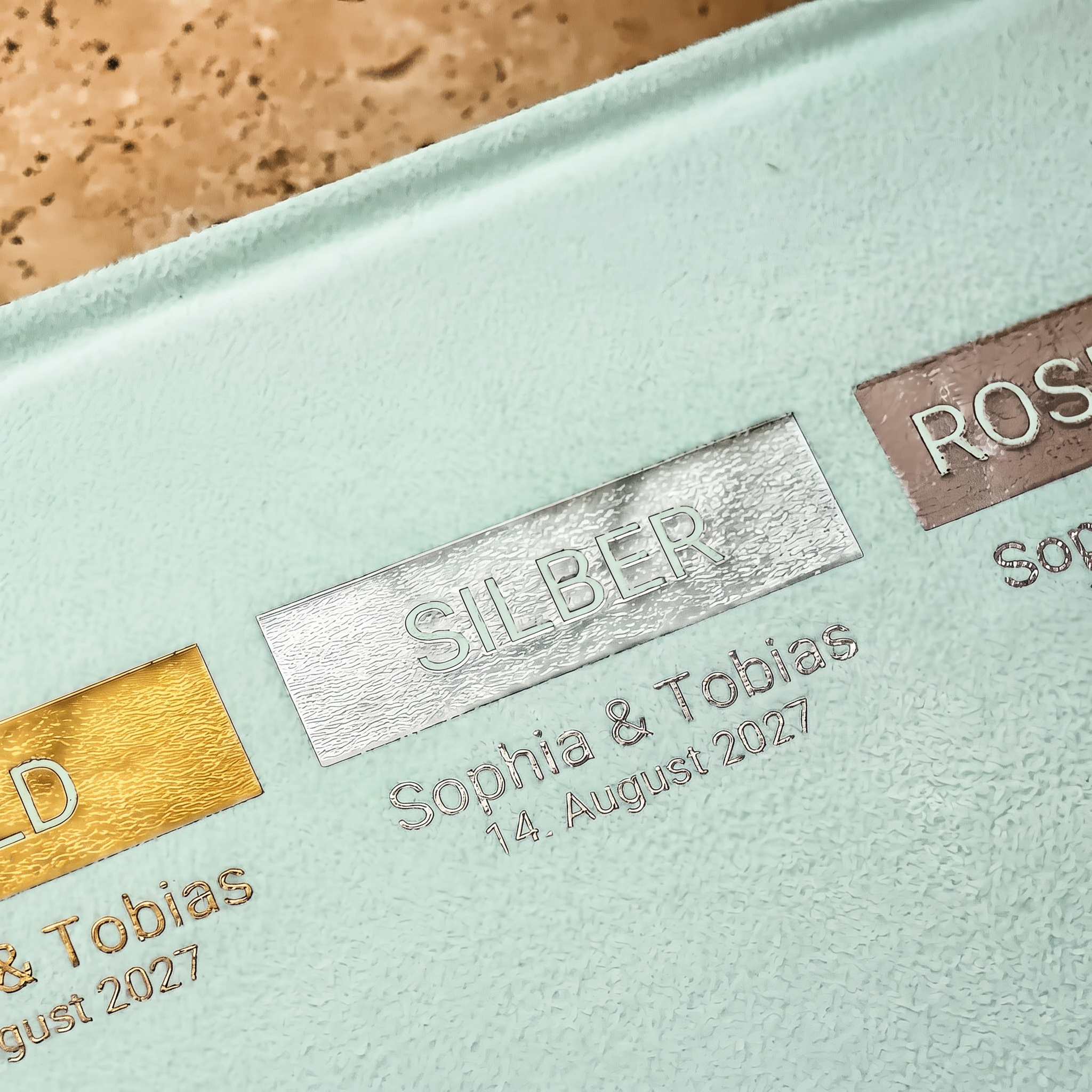 Nahaufnahme einer strukturierten mintgrünen Oberfläche mit geprägtem Metalltext in Gold, Silber und Roségold mit den Bezeichnungen GOLD, SILBER und ROSEGOLD, darunter personalisierte Namen und das Datum 14. August 2027 unter jedem.