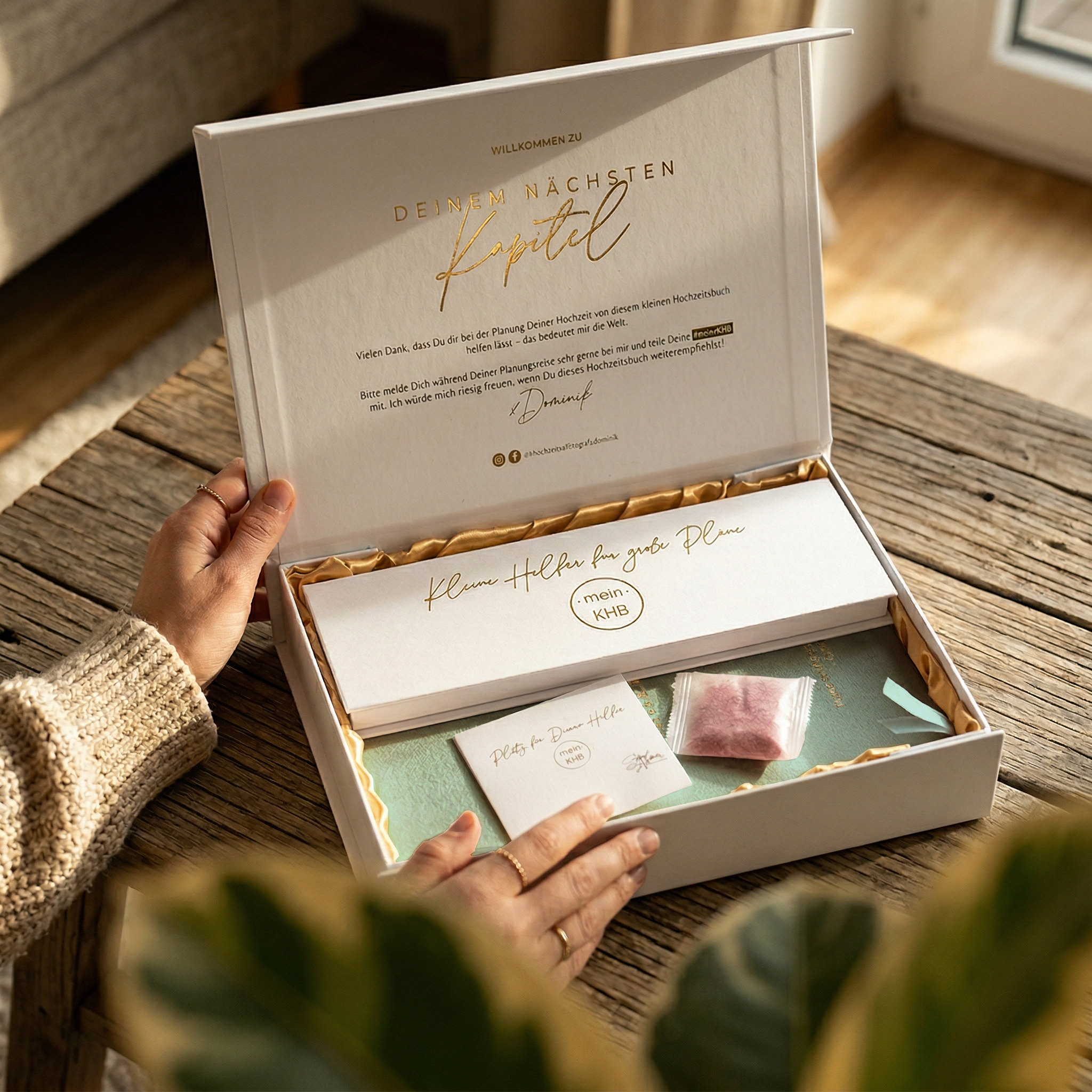 Eine Person öffnet eine weiße Geschenkbox mit deutschem Text auf dem Innendeckel. Die Schachtel enthält ein Büchlein, eine Karte, ein kleines rosafarbenes Tütchen und grünes geschreddertes Papier auf einem Holztisch in der Nähe eines Fensters.