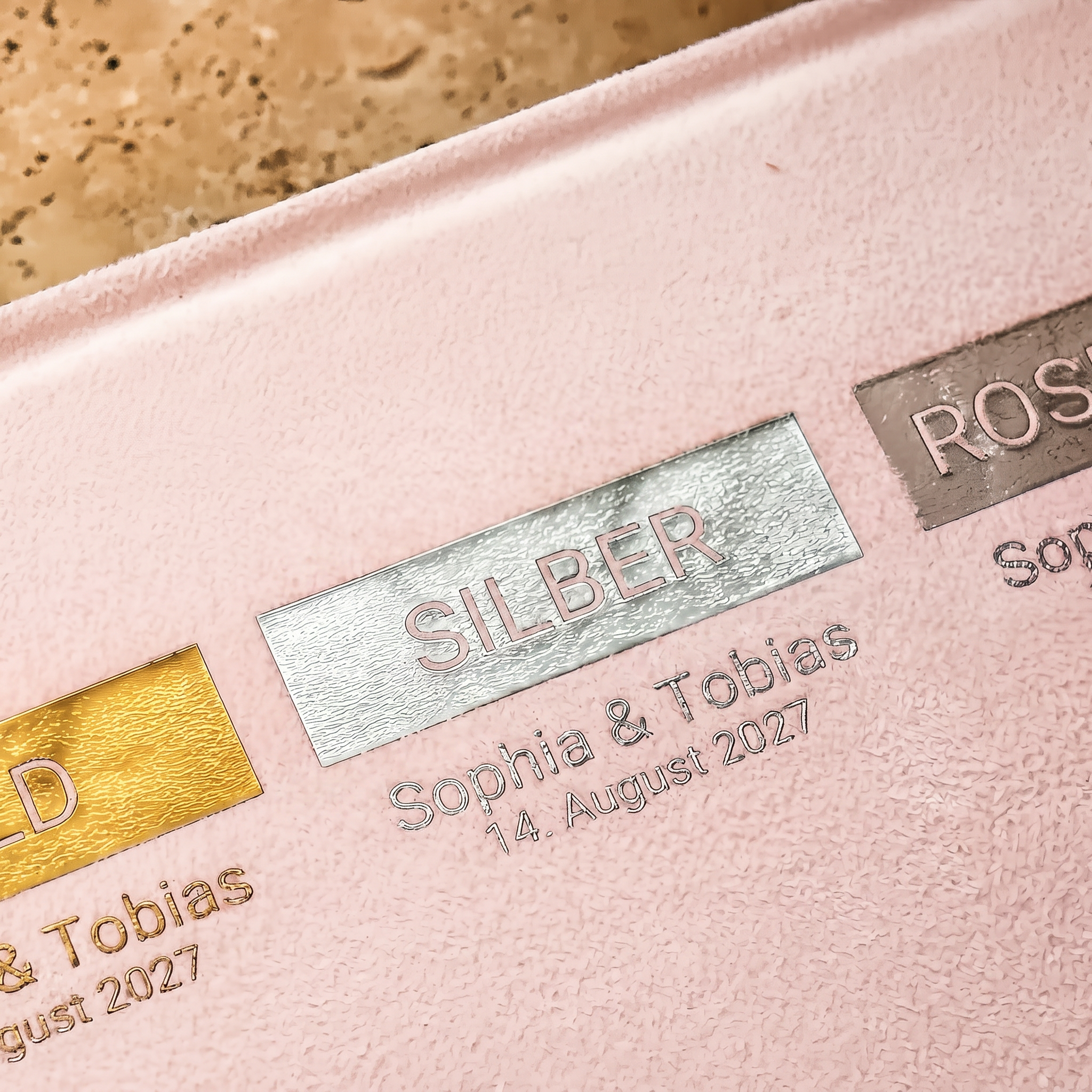 Nahaufnahme von geprägten metallischen Namensschildern auf einer zartrosa Oberfläche, beschriftet mit "GOLD", "SILBER" und "ROSE". Der Text "Sophia  Tobias, 14. August 2027" ist unter dem Schild "SILBER" zu sehen.