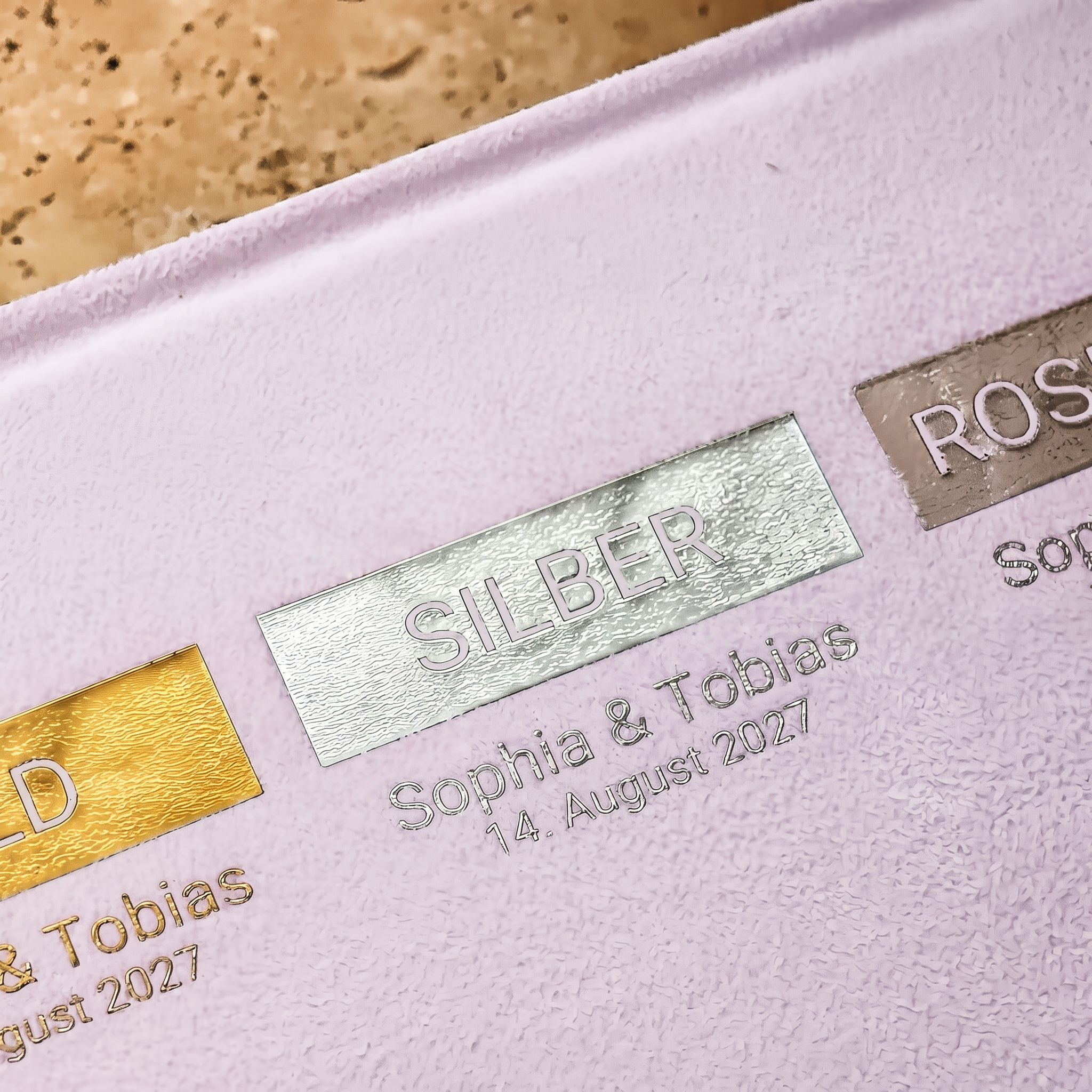 Nahaufnahme einer strukturierten Oberfläche mit geprägten Metallplaketten mit den Bezeichnungen GOLD, SILBER und ROSE. Darunter sind jeweils die Namen Sophia  Tobias und das Datum 14. August 2027 gedruckt.