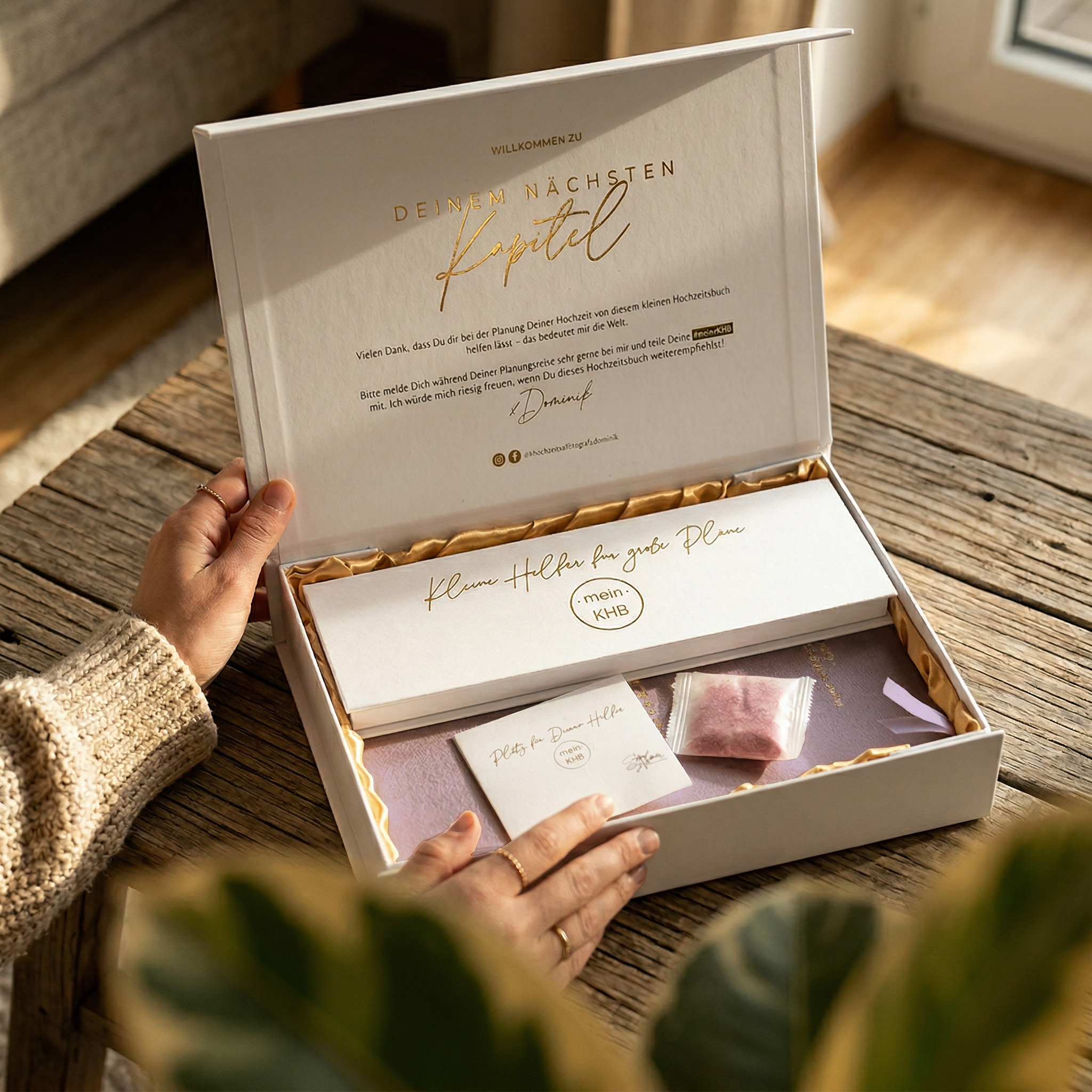 Eine Person öffnet eine weiße Geschenkschachtel mit goldenen Akzenten, die fein säuberlich geordnete Schreibwaren und ein kleines rosafarbenes Tütchen enthält. Auf dem Deckel der Schachtel steht ein Willkommensgruß in deutscher Sprache. Das Sonnenlicht fällt durch ein nahe gelegenes Fenster ein.