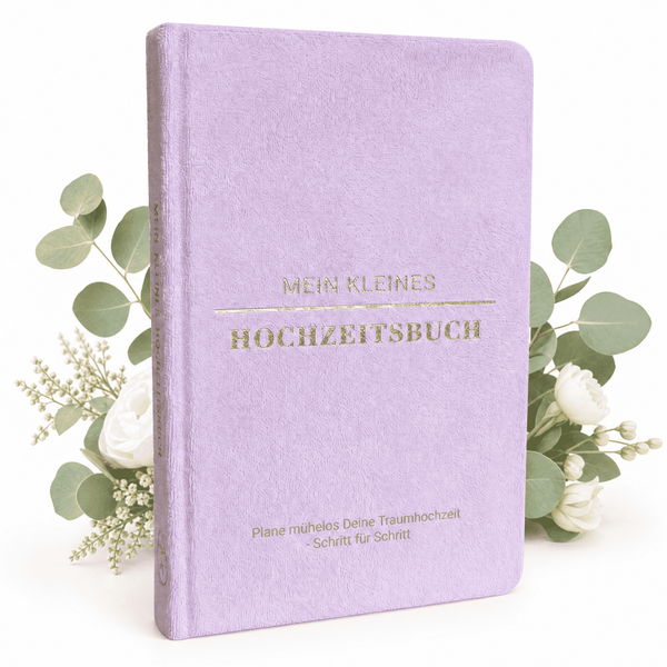 Das pastelllila Mein kleines Hochzeitsbuch - Fürstliche Romantik von Mein kleines Hochzeitsbüro steht aufrecht inmitten von weißen Rosen und Grünzeug. Der goldgeprägte Untertitel lautet: Plane mühelos Deine Traumhochzeit Schritt für Schritt.