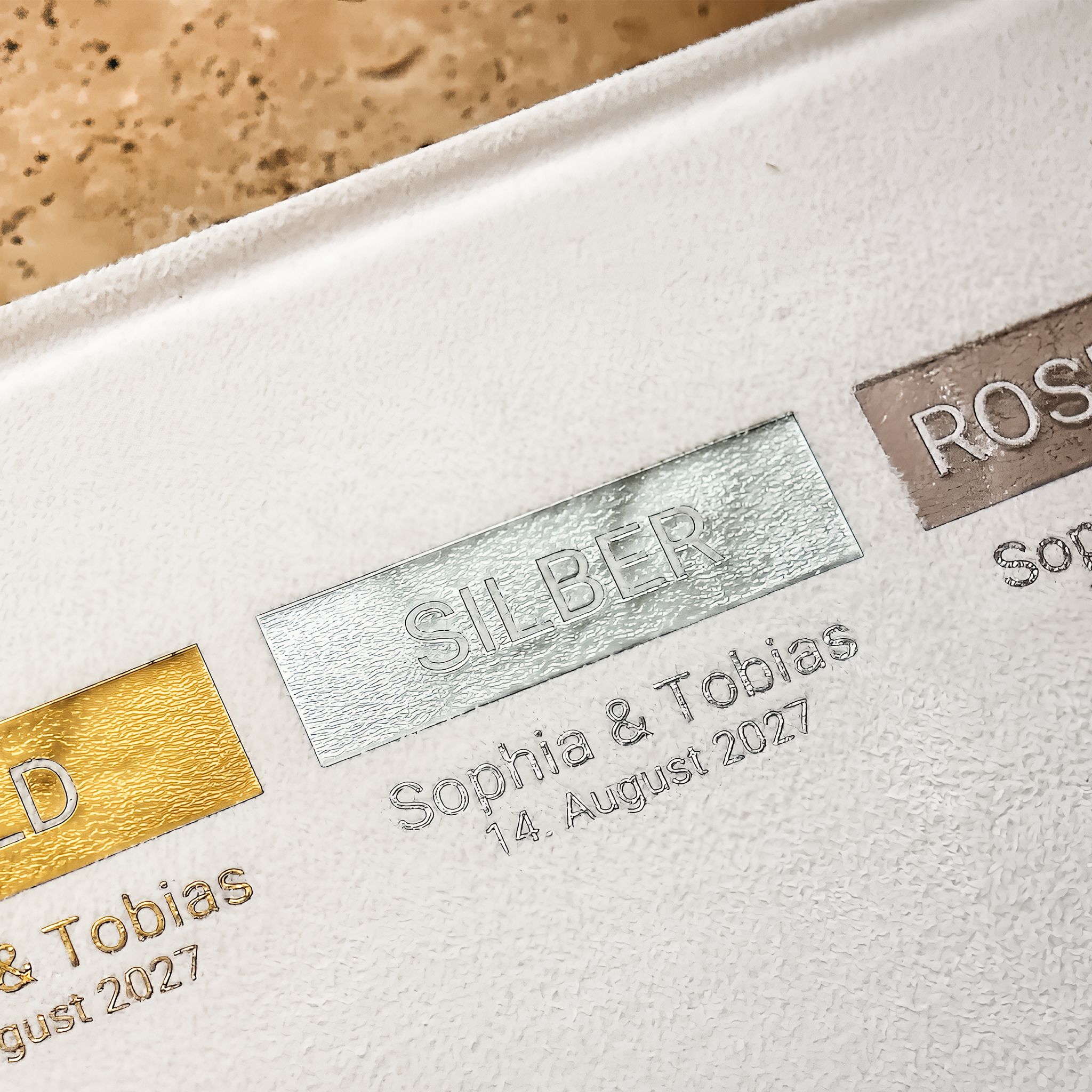 Nahaufnahme einer strukturierten weißen Oberfläche, auf der drei metallische Farbfelder mit den Bezeichnungen GOLD, SILBER und ROSE zu sehen sind, unter denen die Namen Sophia  Tobias und das Datum 14. August 2027 unter jedem gedruckt.