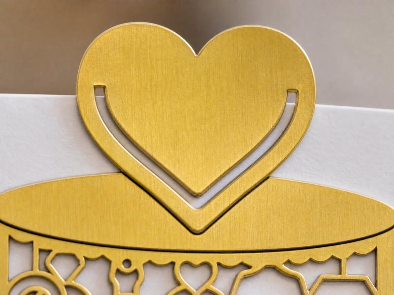 Nahaufnahme einer herzförmigen Dekoration aus Goldmetall, die oben auf einer weißen Karte mit verschlungenen goldenen Mustern angebracht ist.