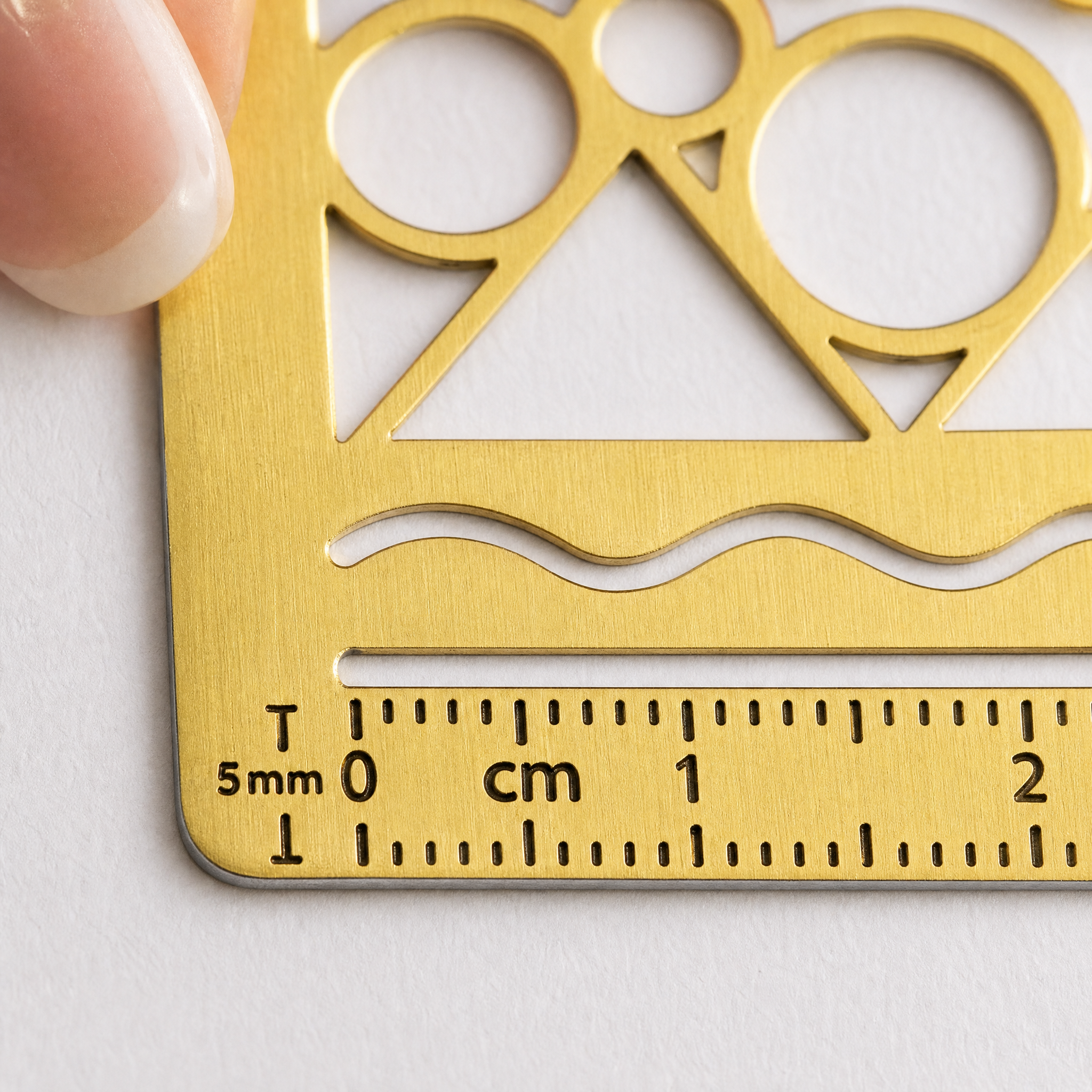 Nahaufnahme einer goldenen Schablone mit geometrischen Formen und einem in Zentimetern und Millimetern markierten Lineal, das von einem Finger an der Ecke gehalten wird.