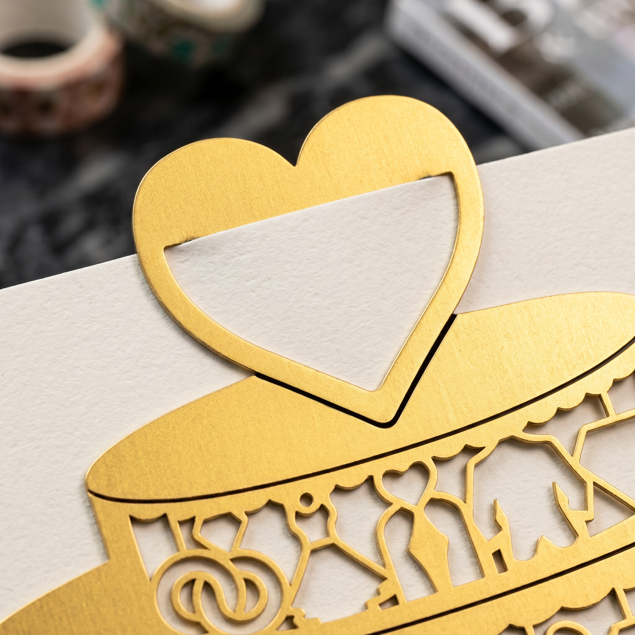 Nahaufnahme einer dekorativen goldenen Papierkarte mit einem herzförmigen Ausschnitt und komplizierten lasergeschnittenen Mustern auf einer weißen Fläche mit unscharfen Objekten im Hintergrund.