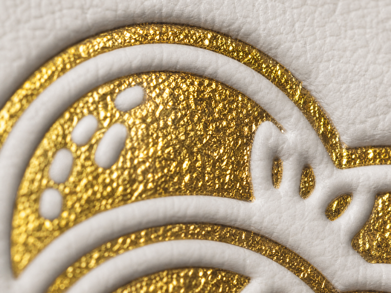 Nahaufnahme eines geprägten Goldfoliendesigns auf strukturiertem weißem Papier, das glänzende, erhabene Goldbereiche und glatte, geschwungene Linien zeigt.