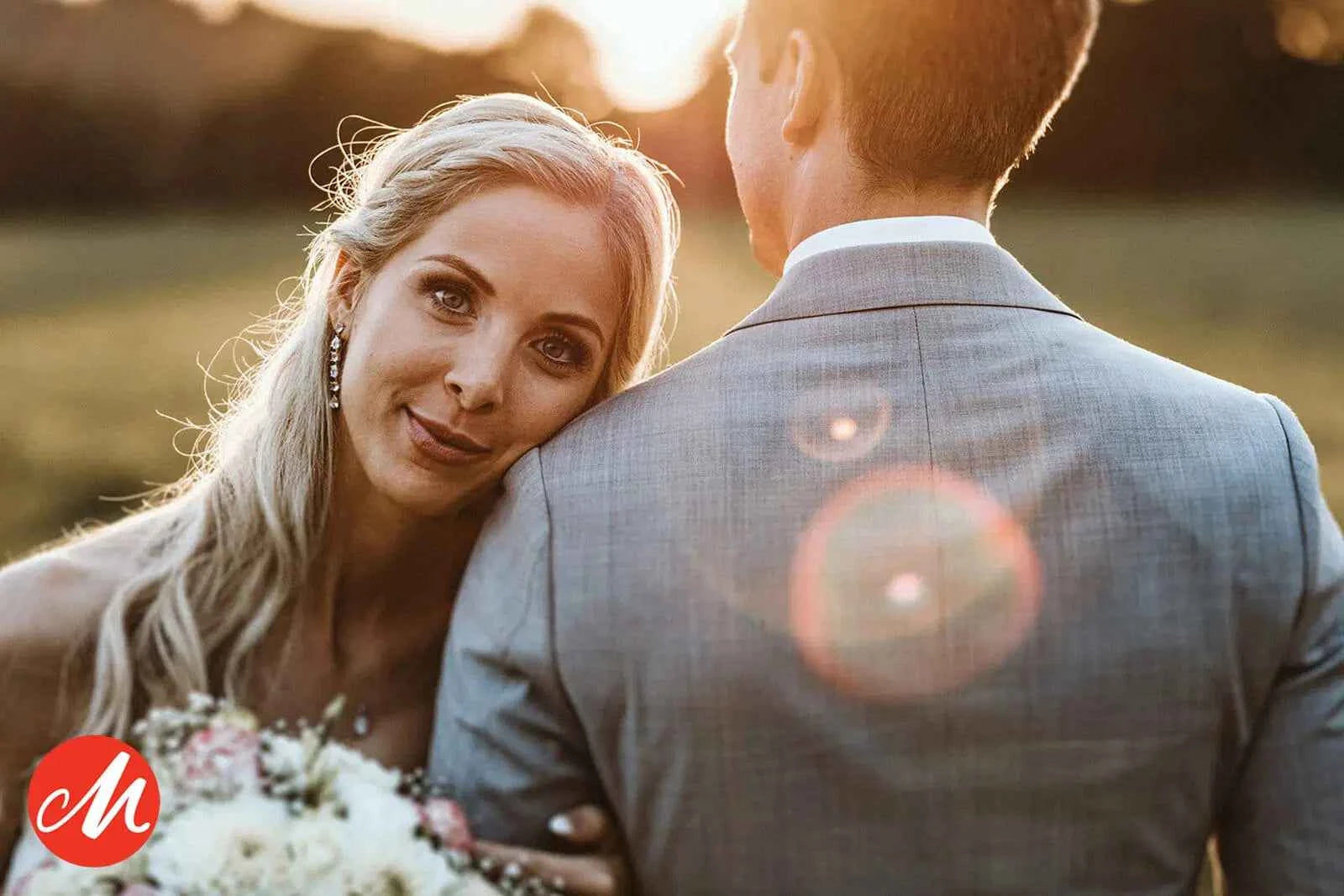 Eine blonde Frau in einem Hochzeitskleid legt ihren Kopf auf die Schulter eines Mannes, der einen Blumenstrauß hält, mit Sonnenlicht und Streulicht im Hintergrund. Der Mann trägt einen grauen Anzug und blickt von der Kamera weg.
