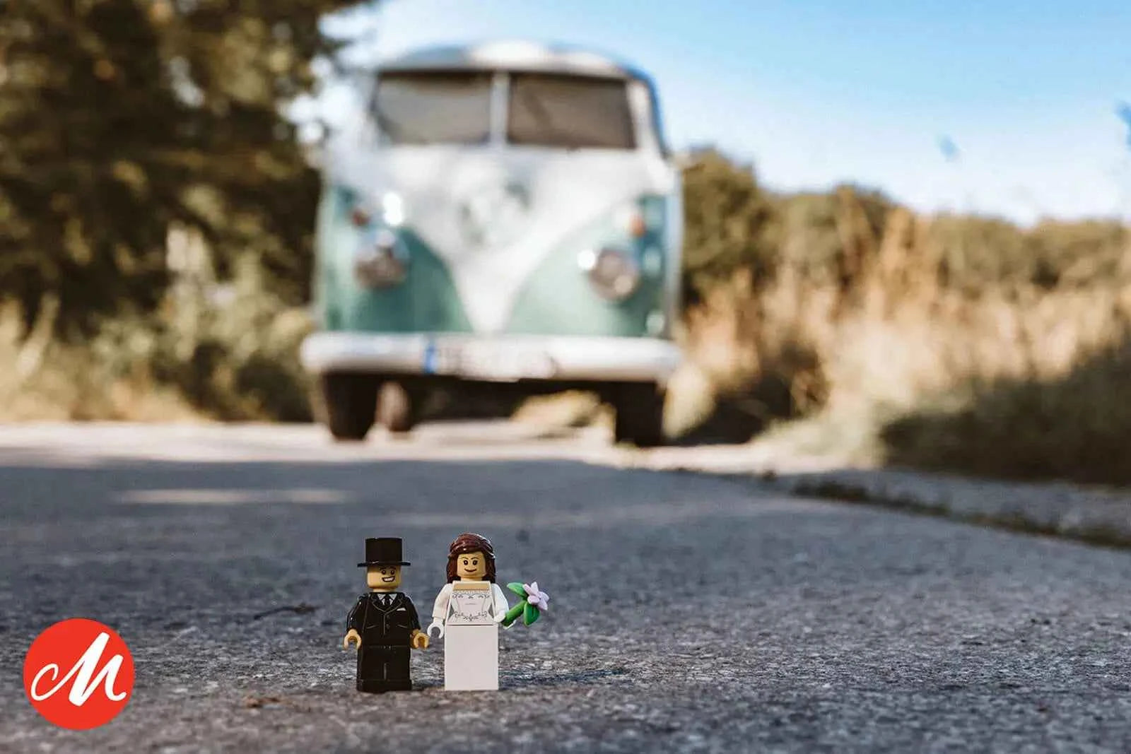 Zwei LEGO Minifiguren, die als Braut und Bräutigam gekleidet sind, stehen an einem sonnigen Tag auf einer Straße, mit einem grünen Lieferwagen im Hintergrund. Ein rotes kreisförmiges Logo befindet sich in der unteren linken Ecke.