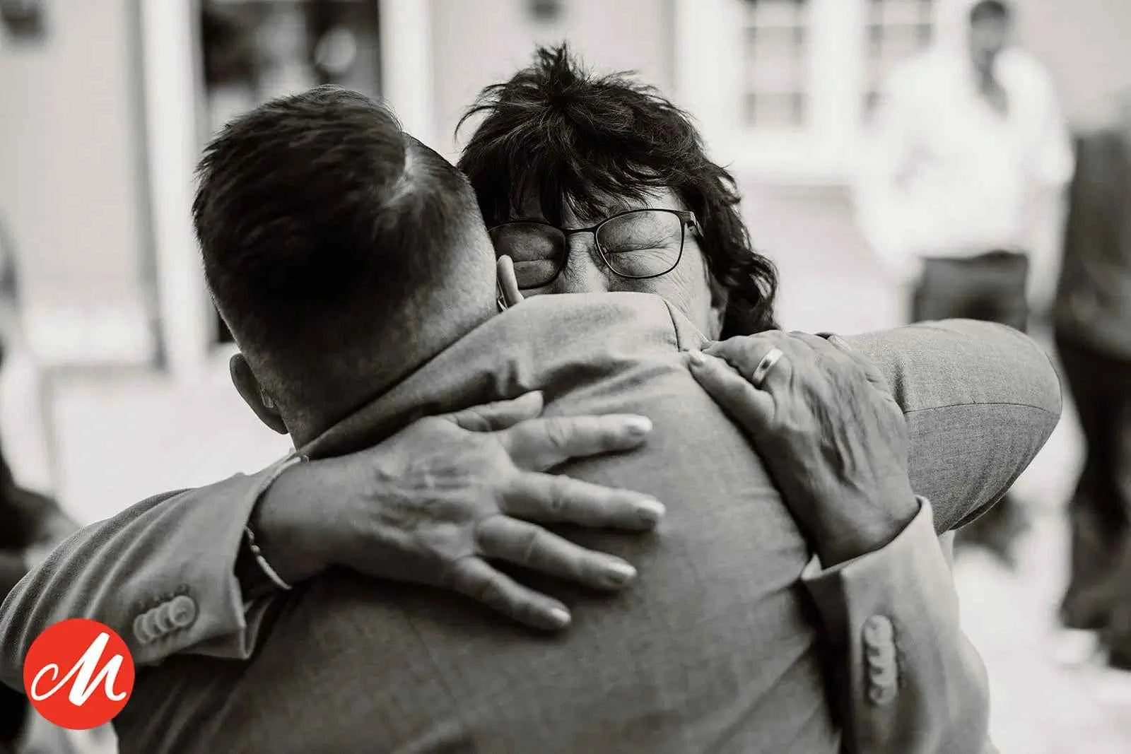 Ein Graustufenfoto von zwei Menschen, die sich eng umarmen. Die eine Person, die eine Brille trägt, wirkt emotional, hat die Augen geschlossen und den Mund leicht geöffnet. Der Rücken der anderen Person ist der Kamera zugewandt. In der unteren linken Ecke befindet sich ein rotes Logo mit dem Buchstaben M.