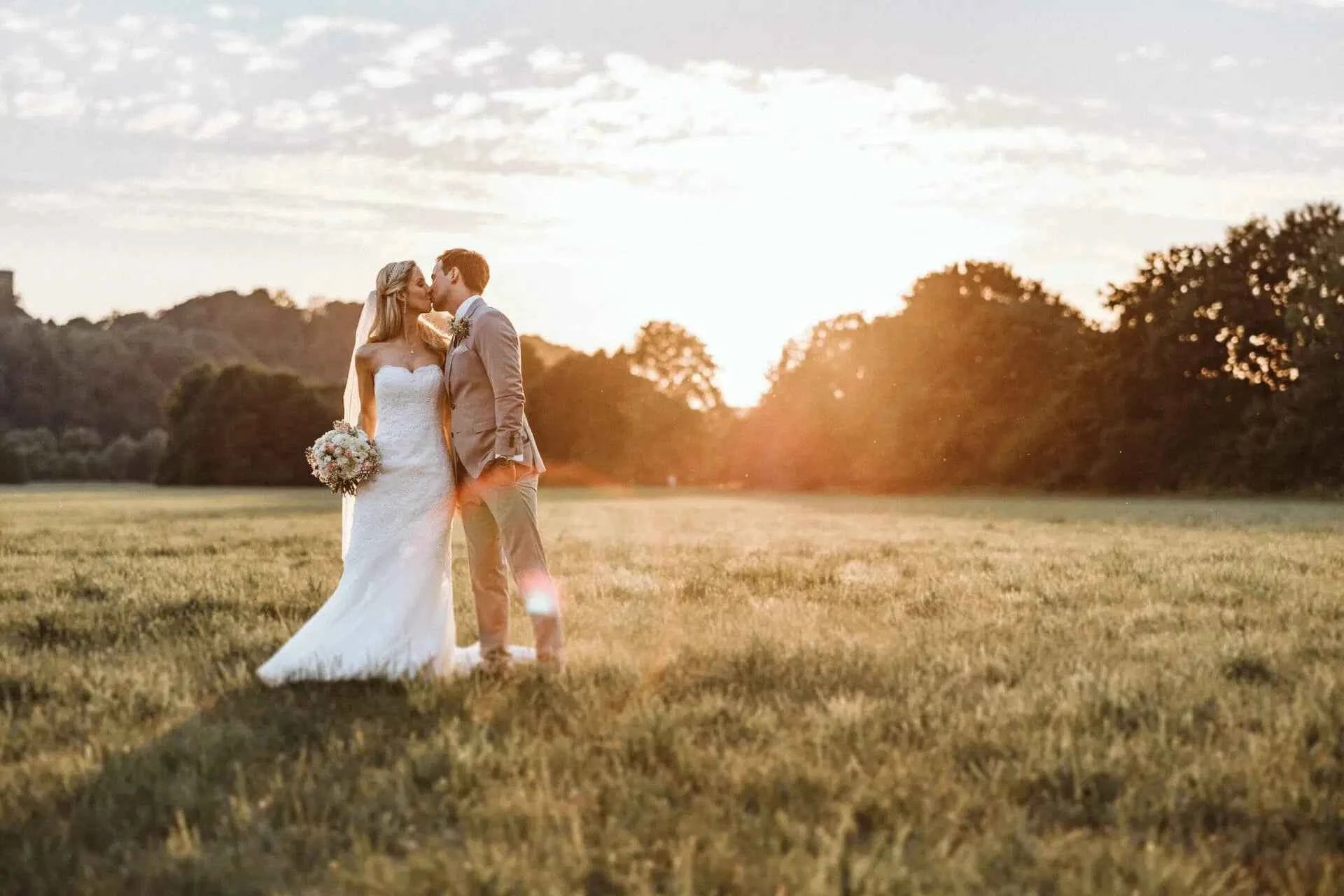 Eine Braut und ein Bräutigam stehen in einem sonnenbeschienenen Feld bei Sonnenuntergang und geben sich einen Kuss. Die Braut trägt ein weißes Kleid und hält einen Blumenstrauß, während der Bräutigam einen hellen Anzug trägt. Im Hintergrund sind Bäume und ein leuchtender Himmel zu sehen.