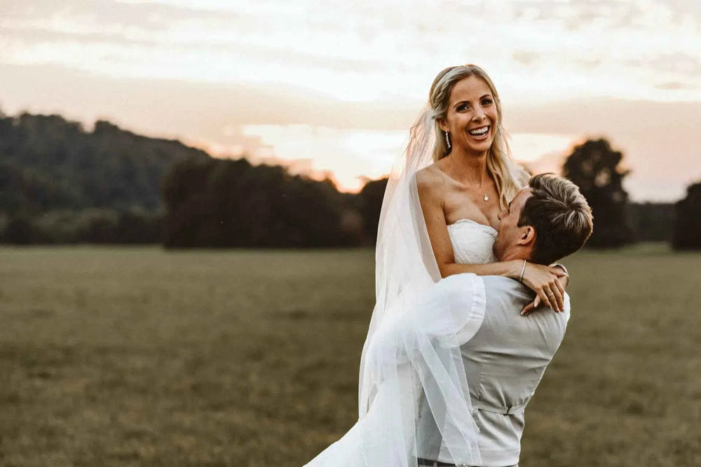Ein Bräutigam hebt eine lächelnde Braut in einem weißen Hochzeitskleid und Schleier hoch, während er in einem grasbewachsenen Feld bei Sonnenuntergang steht, mit Bäumen und Hügeln im Hintergrund.