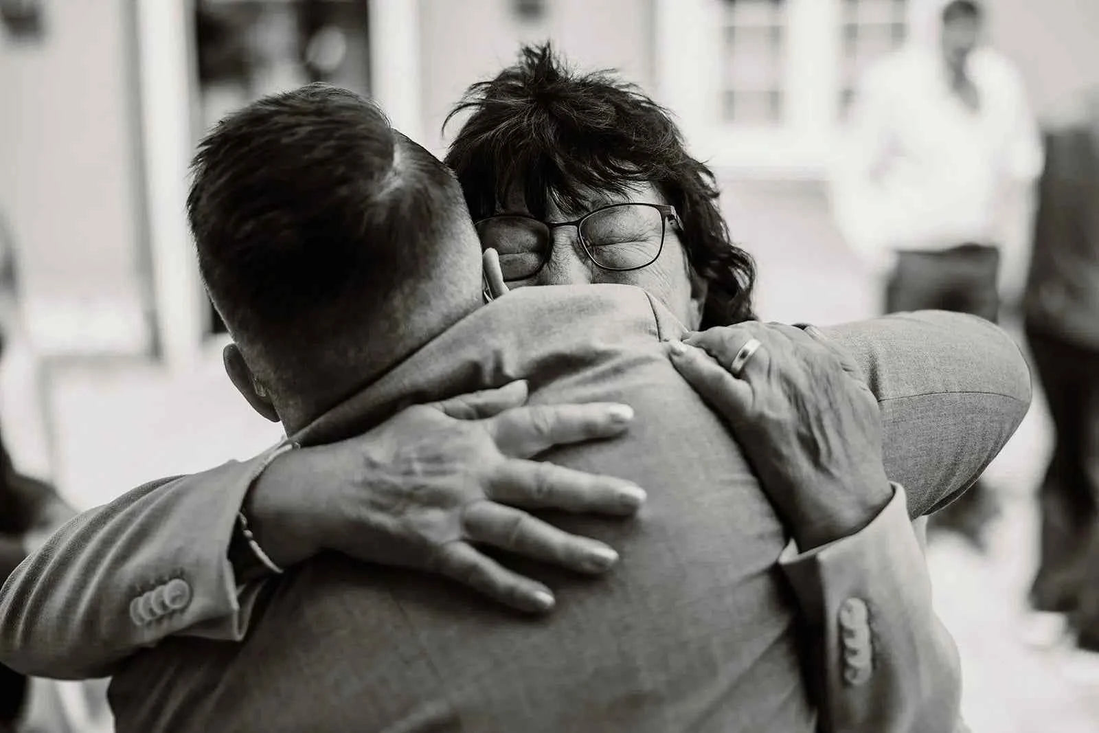 Ein Schwarz-Weiß-Foto von zwei Menschen, die sich eng umarmen, einer mit Brille und dunklem Haar, die Augen in Emotion geschlossen, während der andere sich abwendet und einen Anzug trägt. Der Hintergrund ist sanft verschwommen.