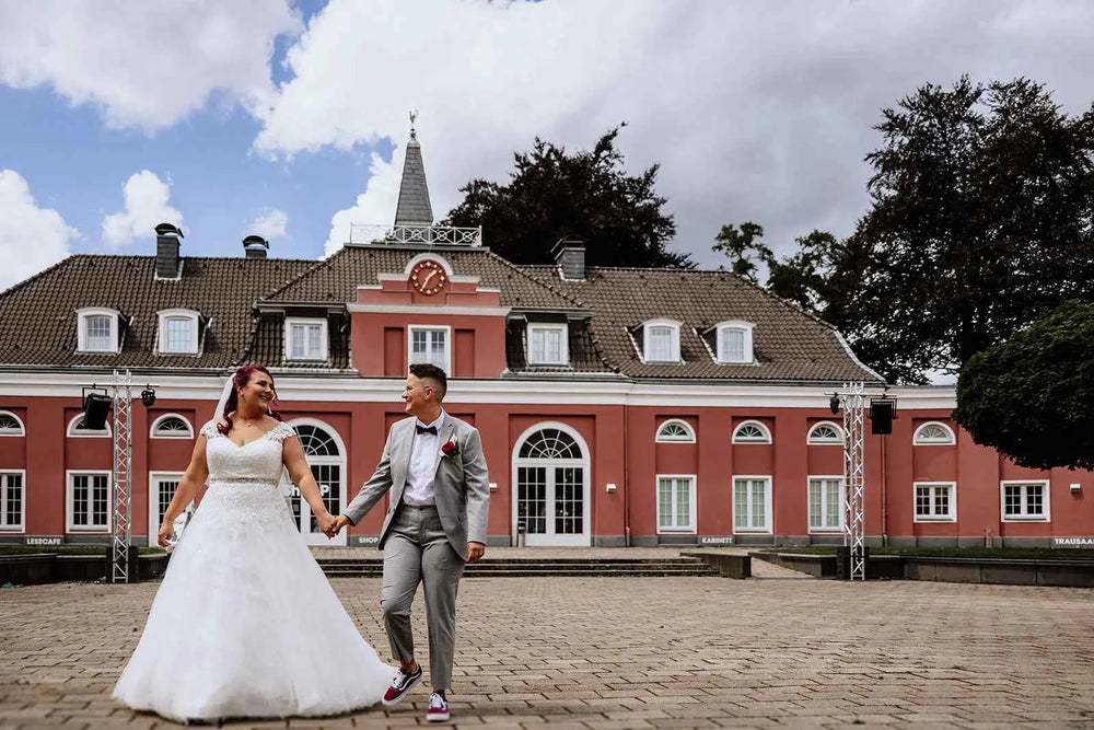 Ein frisch verheiratetes Paar, das sich an den Händen hält und lächelt, steht vor einem großen, eleganten rosafarbenen Gebäude mit weißen Verzierungen und einem Uhrenturm, unter einem teilweise bewölkten Himmel.
