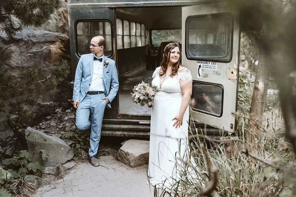 Eine Braut in einem weißen Kleid und mit einem Blumenstrauß in der Hand steht lächelnd vor einem alten Bus, während der Bräutigam in einem hellblauen Anzug am Bus lehnt und zur Seite schaut. Sie sind von Felsen und Grün umgeben.
