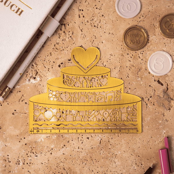 Die Lineal Clip-Schablone "Hochzeitstorte" von Mein kleines Hochzeitsbüro ist eine goldene Metallschablone in Form einer dreistöckigen Hochzeitstorte mit einem Herz-Topper und dekorativen Ausschnitten, die auf einer strukturierten braunen Oberfläche mit Schreibwaren und Münzen präsentiert wird.