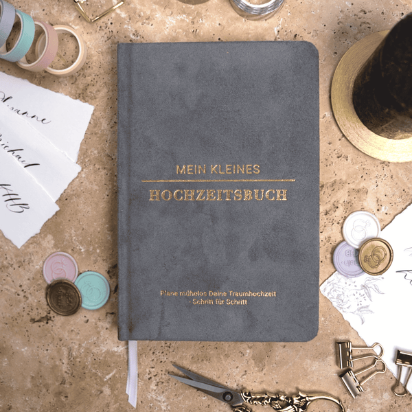 Das graue Mein kleines Hochzeitsbuch - Eleganter Nebel von Mein kleines Hochzeitsbüro, mit Goldprägung, liegt auf Marmor mit Wachssiegeln, Geschenkband, einem Goldstempel und handschriftlichen Notizen - perfekt für eine stilvolle Hochzeitsplanung.