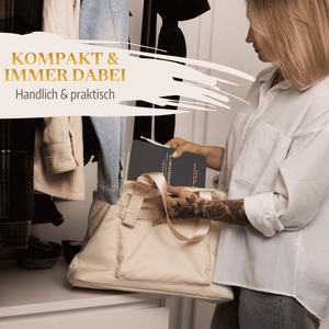 Eine Frau im weißen Hemd legt das schwarze "Mein kleines Hochzeitsbuch - Eleganter Nebel" von Mein kleines Hochzeitsbüro in eine beige Handtasche. Im Hintergrund ist ein Garderobenständer mit Jacken zu sehen. Text auf Deutsch: KOMPAKT  IMMER DABEI, Handlich  praktisch.