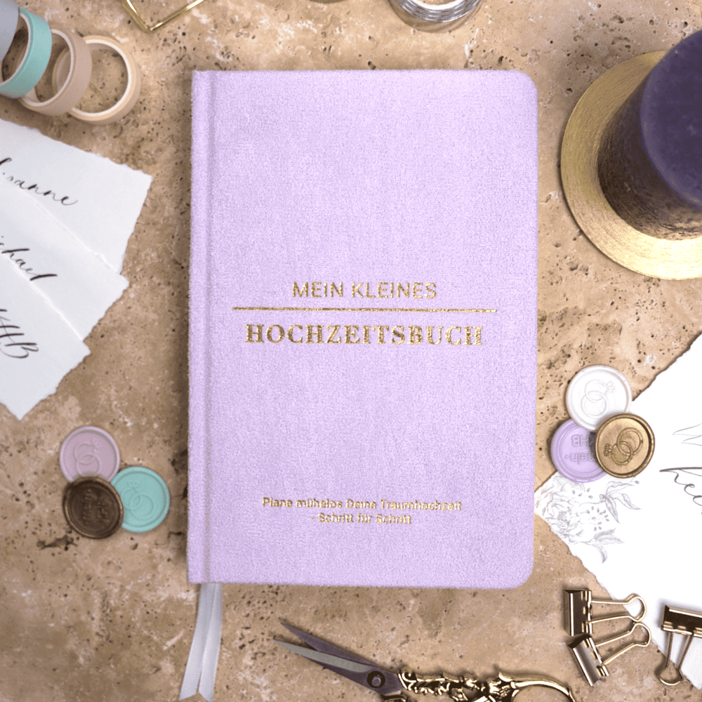 Das pastelllila Mein kleines Hochzeitsbuch - Fürstliche Romantik von Mein kleines Hochzeitsbüro steht aufrecht inmitten von weißen Rosen und Grünzeug. Der goldgeprägte Untertitel lautet: Plane mühelos Deine Traumhochzeit Schritt für Schritt.