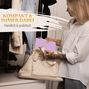Eine Frau in einem weißen Hemd legt "Mein kleines Hochzeitsbuch - Fürstliche Romantik" von Mein kleines Hochzeitsbüro in eine beigefarbene Tasche in ihrem Schrank. In der Nähe hängen Kleidungsstücke. Deutscher Text: KOMPAKT  IMMER DABEI - Handlich  praktisch.