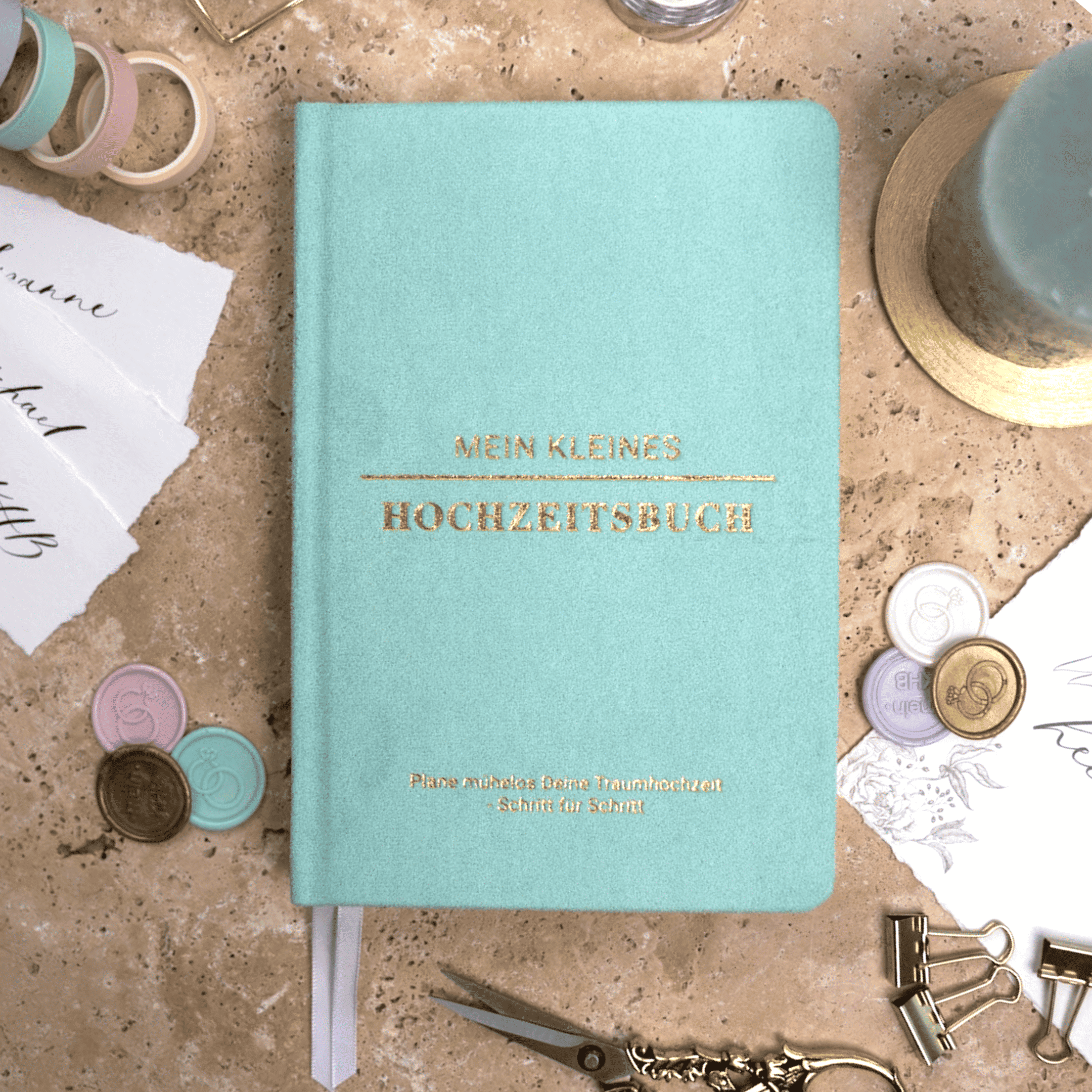 Das hellblaue "Mein kleines Hochzeitsbuch - Zarter Traum" von Mein kleines Hochzeitsbüro ruht auf Marmor inmitten von Briefpapier, Washi-Tapes, goldenen Wachssiegeln, Clips und einer blauen Kerze - der perfekte Planungsbegleiter für die Hochzeitsplanung.