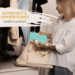 Eine Frau legt das türkisfarbene Mein kleines Hochzeitsbuch - Zarter Traum von Mein kleines Hochzeitsbüro in eine cremefarbene Tragetasche in einem Raum mit hängender Kleidung. Text: KOMPAKT  IMMER DABEI - Dein praktischer Planungshelfer für die Hochzeitsplanung.
