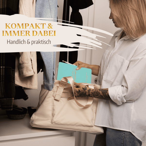 Eine Frau legt das türkisfarbene Mein kleines Hochzeitsbuch - Zarter Traum von Mein kleines Hochzeitsbüro in eine cremefarbene Tragetasche in einem Raum mit hängender Kleidung. Text: KOMPAKT  IMMER DABEI - Dein praktischer Planungshelfer für die Hochzeitsplanung.