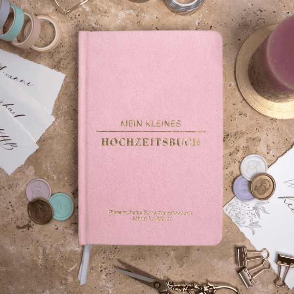 Ein rosafarbenes Mein kleines Hochzeitsbuch - Zuckerwatten Herzchen von Mein kleines Hochzeitsbüro ruht auf Marmor, umgeben von Wachssiegeln, Schleife, Kerze, Stift, Papierschnipseln und pastellfarbenen Klammern - ideal für Hochzeitsplanung und Checklisten.
