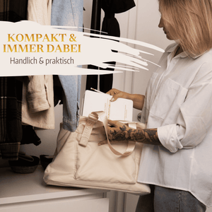 Eine Frau in einem weißen Hemd legt "Mein kleines Hochzeitsbuch - Ewige Eleganz" von Mein kleines Hochzeitsbüro in eine beigefarbene Tasche in einer Halle mit Mänteln. Deutscher Text: "Kompakt  immer dabei. Handlich  praktisch.