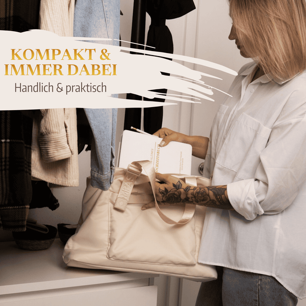 Eine Frau in einem weißen Hemd legt "Mein kleines Hochzeitsbuch - Ewige Eleganz" von Mein kleines Hochzeitsbüro in eine beigefarbene Tasche in einer Halle mit Mänteln. Deutscher Text: "Kompakt  immer dabei. Handlich  praktisch.