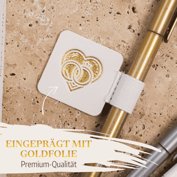 Der Stiftehalter - Schlaufe Extragroß von Mein kleines Hochzeitsbüro ist quadratisch, weiß und ziert zwei goldgeprägte Herzen. Befestigt an einem goldenen Stift auf beigem Steinsockel - Premium-Qualität für die Hochzeit.