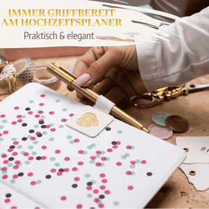 Eine Hand steckt einen goldenen Stift in die Stiftehalter-Schlaufe Extragroß von Mein kleines Hochzeitsbüro am Notizbuch mit bunten Konfetti-Punkten. Goldene Accessoires und Wachssiegel ergänzen den eleganten Organizer für Hochzeiten.