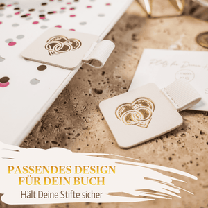 Zwei "Stiftehalter - Schlaufe Standard" von Mein kleines Hochzeitsbüro mit goldenen Ringen liegen auf gesprenkelter Fläche neben einem Notizbuch. Perfekt als Hochzeits-Helfer im passenden Design zu deinem Buch.