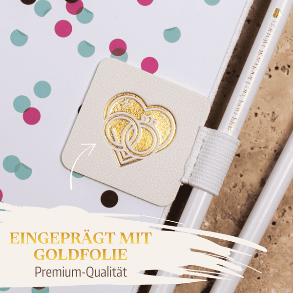 Nahaufnahme der Stiftehalter - Schlaufe Standard von Mein kleines Hochzeitsbüro, ein weißes Notizbuch mit bunten Punkten und Goldfolienherz an der Schließe. Deutscher Text: "Eingeprägt mit Goldfolie - Premium-Qualität." Neben dem Notizbuch liegt ein weißer Stift bei.