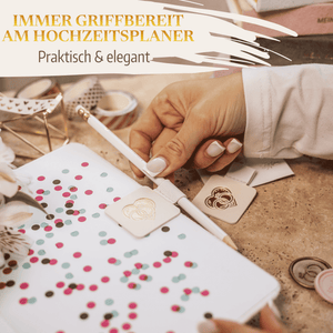 Eine Hand schreibt in ein Hochzeitsplaner-Notizbuch mit einem weißen Stift aus dem Mein kleines Hochzeitsbüro Stiftehalter - Schlaufe Standard. Buntes Konfetti, Anhänger, Klebeband und Blumen schmücken den beigen Schreibtisch. Text: Immer griffbereit am Hochzeitsplaner.